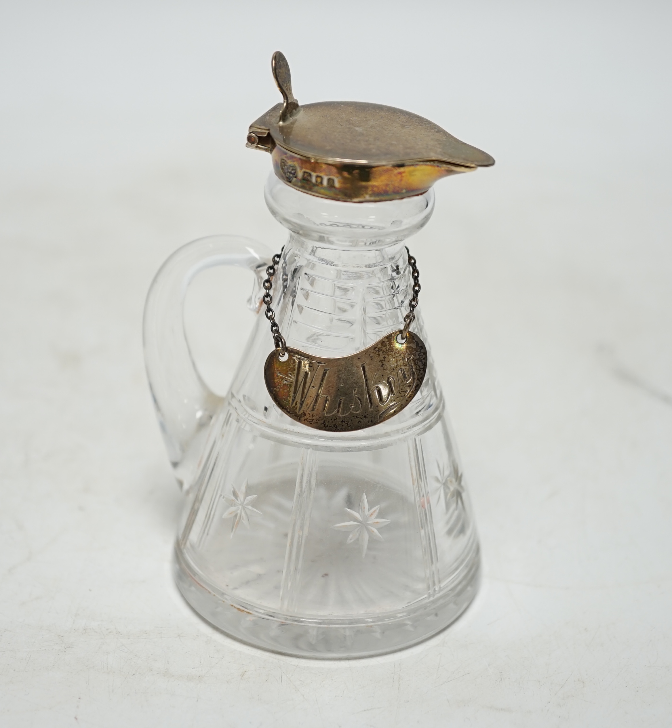 A George V silver mounted cut glass whisky noggin, Goldsmiths & Silversmiths Co Ltd, London, 1933, 10.9cm, with an Edwardian silver 'Whisky' wine label. Condition - poor